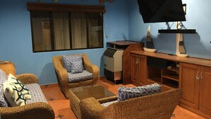 A living room with wicker furniture, a flat-screen TV, and a window with blinds. -  Beach Front House in Tránsito Beach , 5-Bedrooms, AC ,with Beachfront  Pool. (El Tránsito)