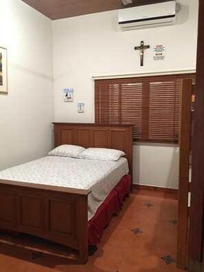Un dormitorio con cama de madera, falda de cama colorada, pared blanca, ventana con persianas y unidad de aire acondicionado.