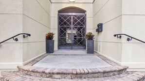 Une entrée d’immeuble avec une porte grillagée, deux plantes en pot et un chemin de briques menant à la porte.