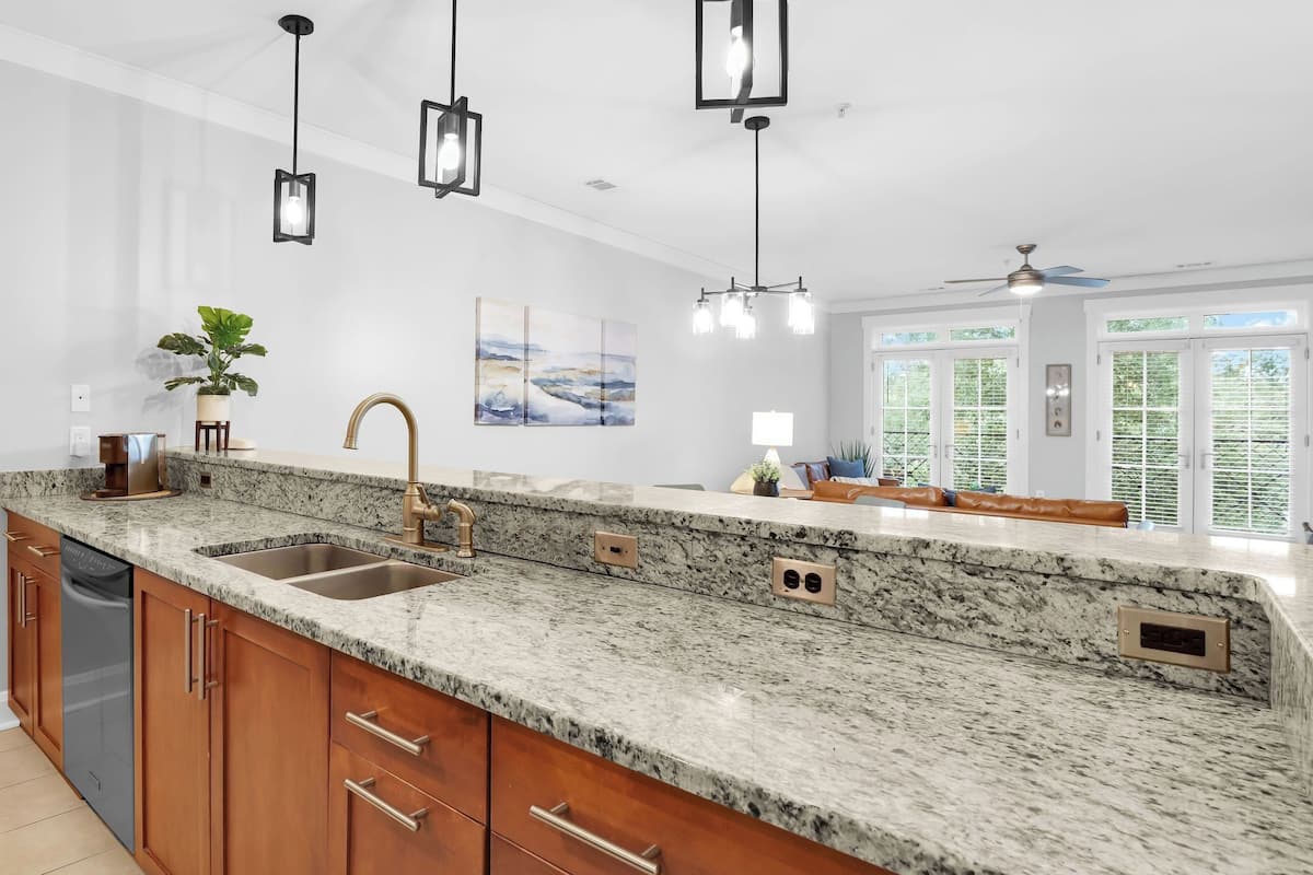 A kitchen with granite countertops, a stainless steel sink, wooden cabinets, pendant lights, and a view to the outside.