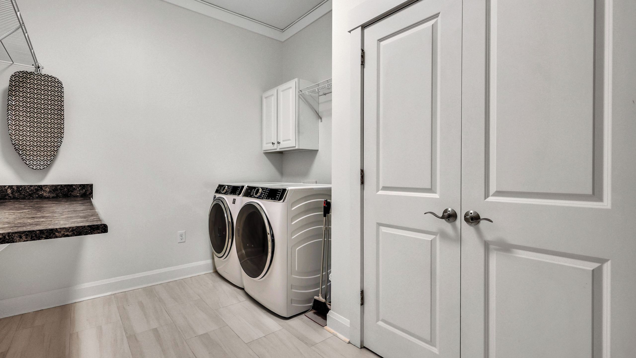 A laundry room with two front-loading washing machines, a metal drying rack, a white door, and a white cabinet above the machines.