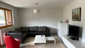 A modern living room with a sectional sofa, a red chair, a white coffee table, a flat-screen TV on a white stand, and a painting on the wall. - Very comfortable 3-room apartment for 3-4 persons : Living room - kitchen with dishwasher, filter co (Grimentz)