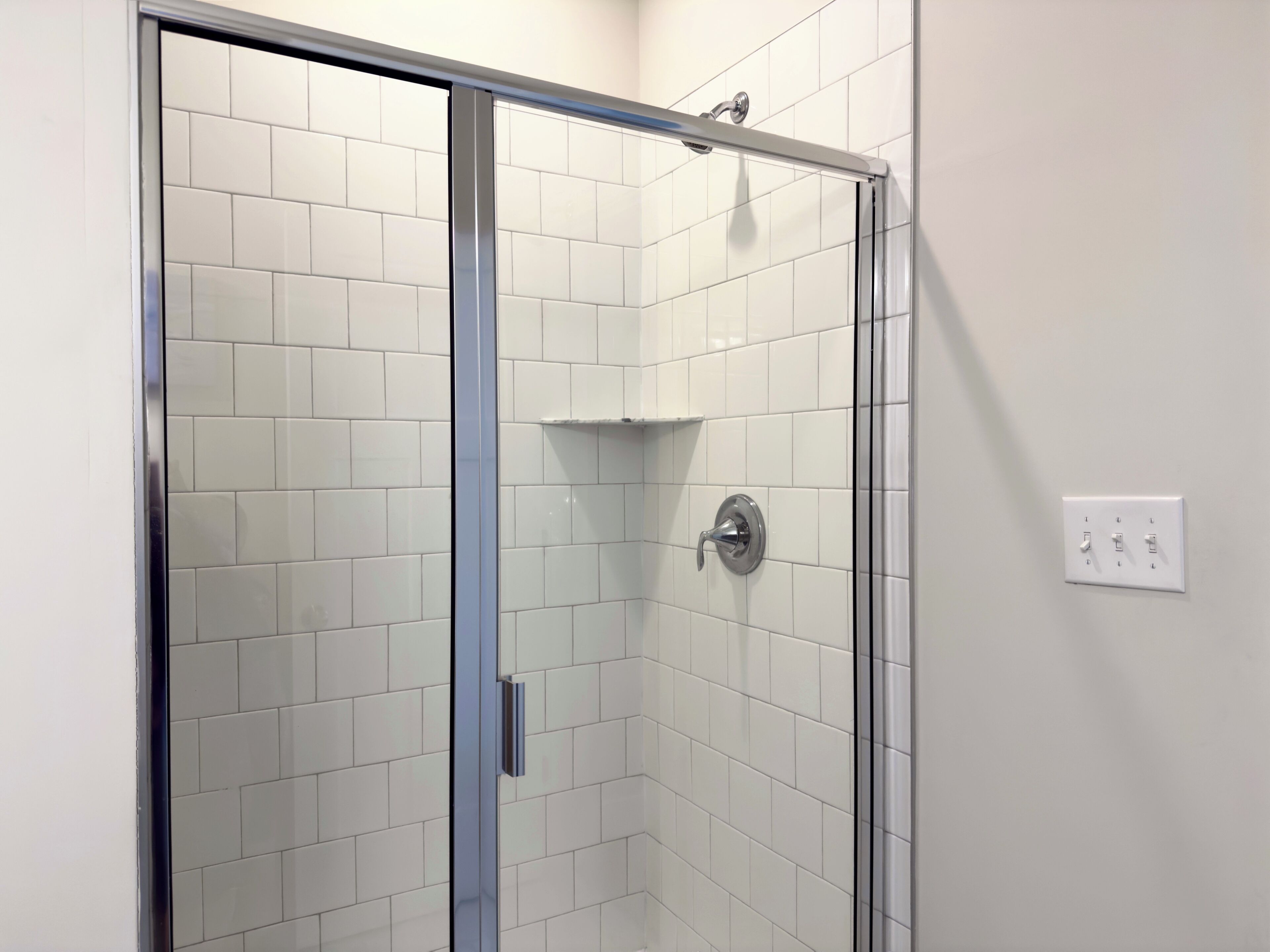 Eine Dusche mit Glastür, weißen Fliesen und einer wandmontierten Leuchte.