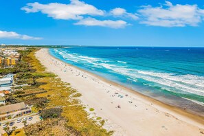 A beach with a clear view of the ocean, sandy shore, and a few buildings in the background. - Oasis by the Sea — Stylish Dog-Friendly Escape with Private Pool, Grill, & Outdoor Bar! 🍹 (Cape Canaveral)