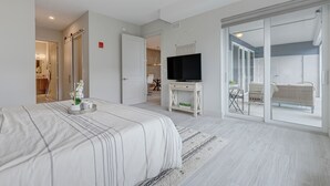 A bedroom with a bed, a TV on a stand, a sliding glass door to a balcony, and a ceiling fan. - Luxurious 4BR Ocean‑View Retreat with Soaking Tub & Pool Access 🌊 (Cocoa Beach)
