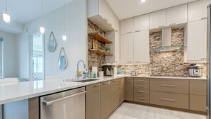 A modern kitchen with a dishwasher, stainless steel appliances, wooden shelves, and a stone backsplash. - Luxurious 4BR Ocean‑View Retreat with Soaking Tub & Pool Access 🌊 (Cocoa Beach)
