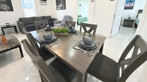 Een eethoek met een houten tafel voor vier personen, omringd door stoelen. Er is een klein bijzettafeltje met een lamp, een plafondventilator en een raam met jaloezieën.