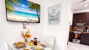 A dining area with a table set for breakfast, a TV mounted on the wall, and a kitchen in the background. - Hollywood Beach 1BR | 30 Sec to Sand + Kitchen (Hollywood)