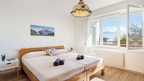 A bedroom with a wooden bed, a bedside table, a nightstand, a window with a view of mountains, and a hanging light fixture. - Cosy apartment for 4 people with WIFI, TV, balcony and panoramic view (Bluche)
