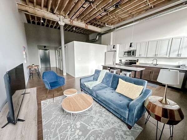 A modern living room with a blue sofa, a coffee table, and a TV. There is a kitchen area with white cabinets and a dining table. - Sleepover | Spacious 1BD/1BA Loft - Newton (Newton)