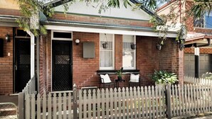 A residential house with a brick facade, a white picket fence, and a small porch area with a table and chairs. - Charming 2Bed Home Near Cafés & Racecourse (Hamilton)
