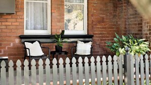A brick building with a white picket fence, two black wicker chairs with white cushions, a small table, and hanging planters. - Charming 2Bed Home Near Cafés & Racecourse (Hamilton)