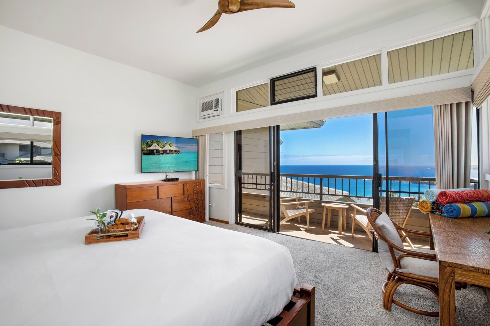 A hotel room with a bed, a TV, a wooden dresser, a balcony with a view of the ocean, and a dining area with chairs and a table.