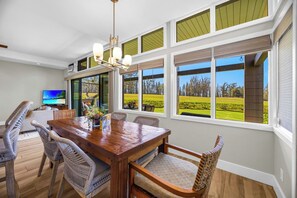 A dining area with a wooden table and wicker chairs, large windows with blinds, and a television on the left. - Kapalua Ridge | Ocean View 5 BR, Sleeps 18 | Car Incl. w/ 6+ Nights | KRV ML-2513 by KBM (Lahaina)