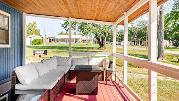 A covered outdoor seating area with a sofa and table, a red carpet, and a view of a grassy yard and buildings in the background. - Bixby Cove Cabin #1 (Porum)