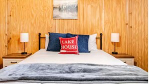 A bedroom with a wooden wall, a bed with a white comforter, two bedside tables with lamps, and a pillow with "LAKE HOUSE" text. - Bixby Cove Cabin #1 (Porum)