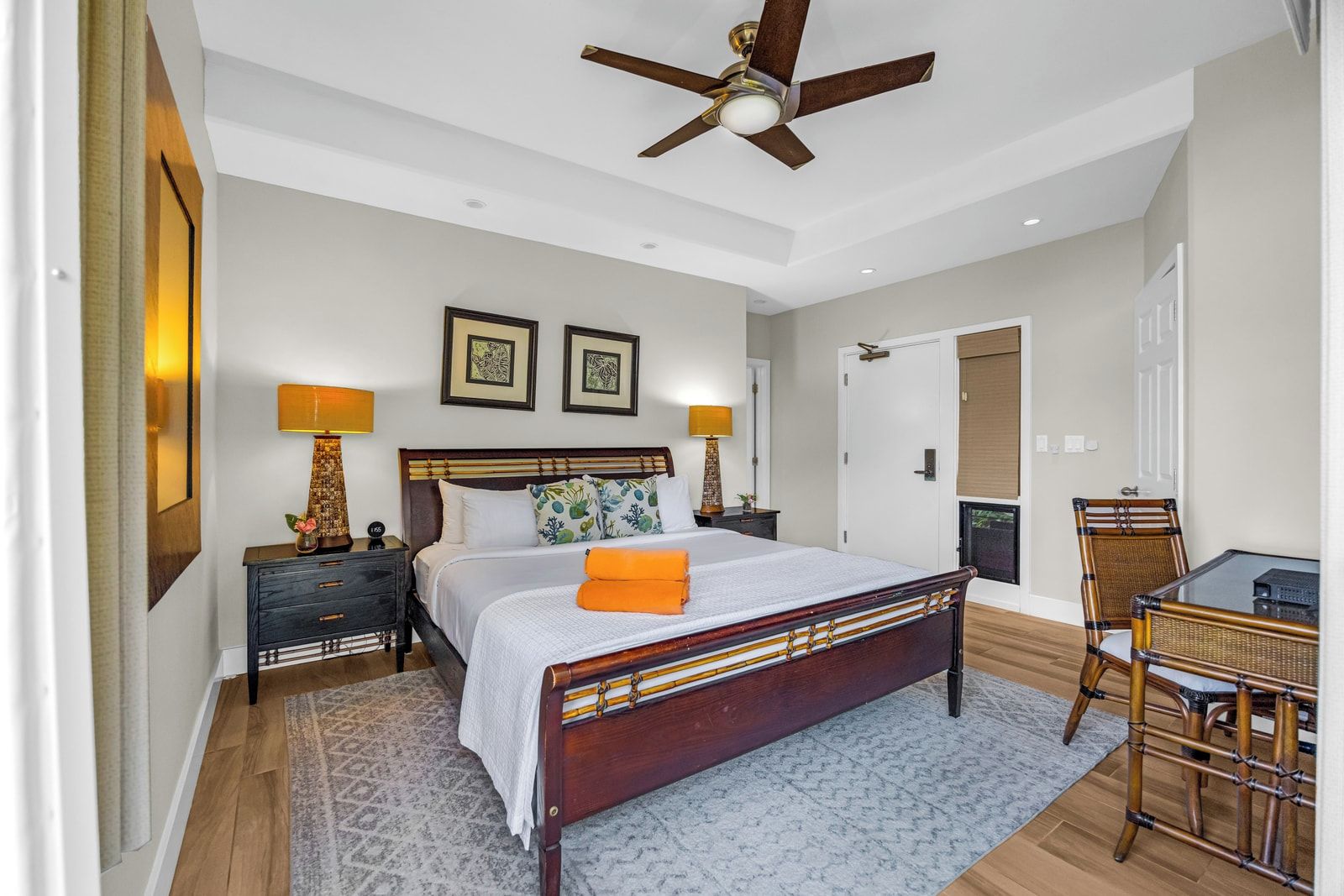 A bedroom with a bed, bedside tables, a ceiling fan, framed artwork, and a door.