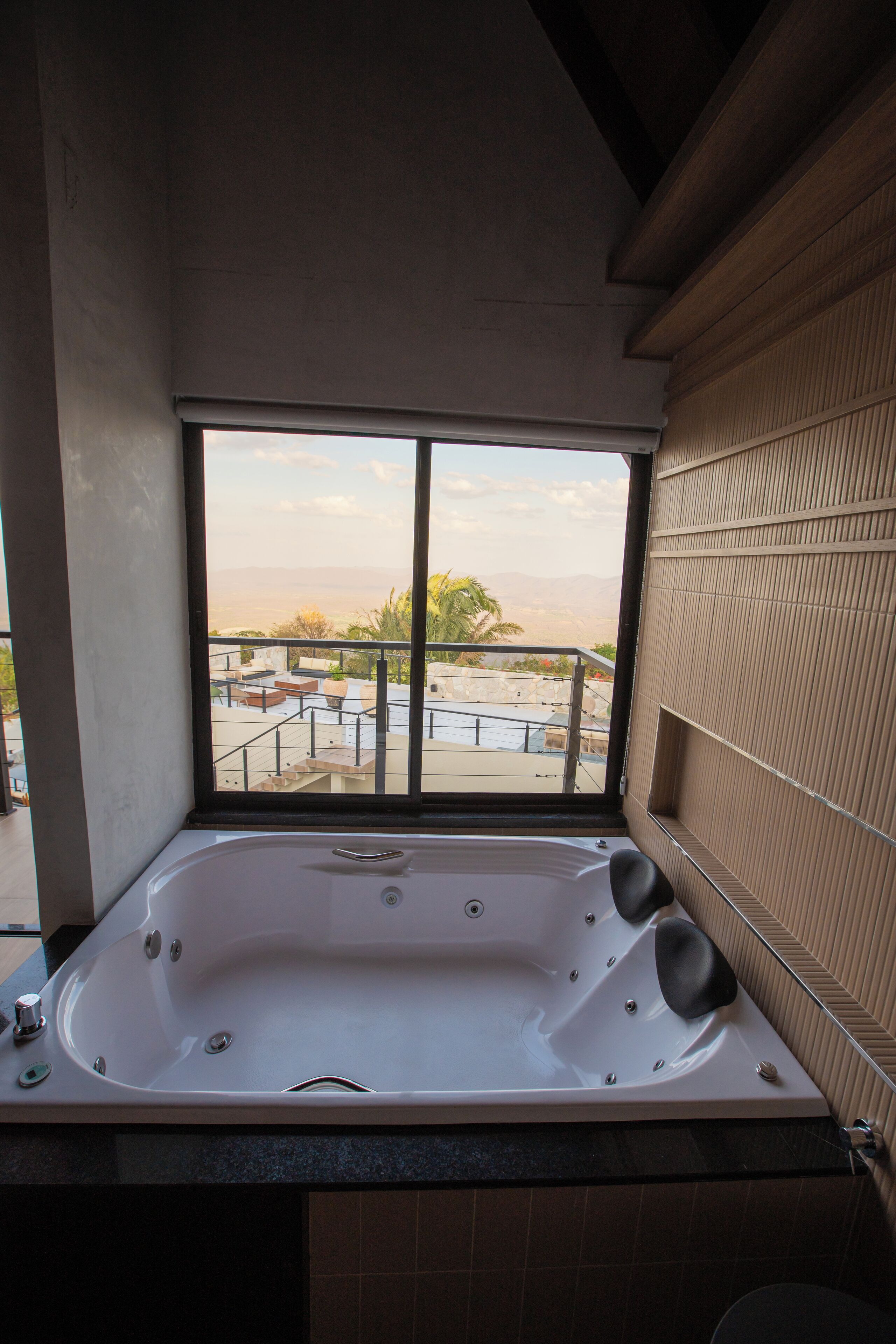 A bathroom with a large bathtub, a window with a view of a balcony and palm trees, and tiled walls.