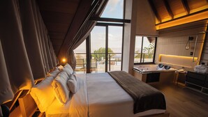 A modern hotel room with a large bed, a desk, and a view of the cityscape through large windows. - Villa Real Chalés (Viçosa do Ceará)