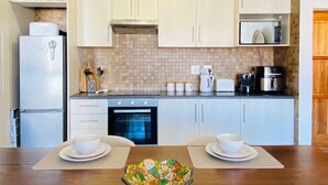 Une cuisine moderne avec des meubles blancs, un four à micro-ondes, une cuisinière et un réfrigérateur. On y trouve une table à manger avec deux assiettes blanches et un saladier coloré au centre.