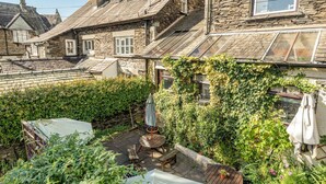 A stone building with a sloped roof, a wooden deck with outdoor furniture, and a garden area with various plants and flowers. - Ambleside Guest House (Ambleside)
