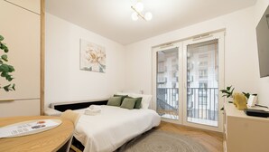 A modern hotel room with a bed, a desk, a radiator, a window with blinds, and a plant. - Posnania Deluxe Apartment (Poznań)