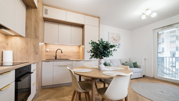 A modern kitchen with wooden cabinets, a dining table with chairs, a potted plant, and a view of the city through a large window. - Posnania Deluxe Apartment (Poznań)