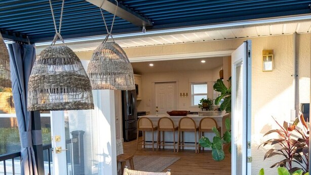 Un espace extérieur aménagé pour les repas, avec une table et des chaises en bois, une terrasse couverte avec un toit bleu, des lampes en osier suspendues et une vue sur l’intérieur de la maison.