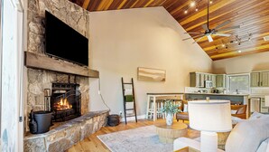 A spacious living room with a stone fireplace, a television mounted on the wall, a ceiling fan, and a kitchen area with green cabinets. - NEW! Ski-In/Out, Lone Peak & Lake Views, 3B/3B (Big Sky)