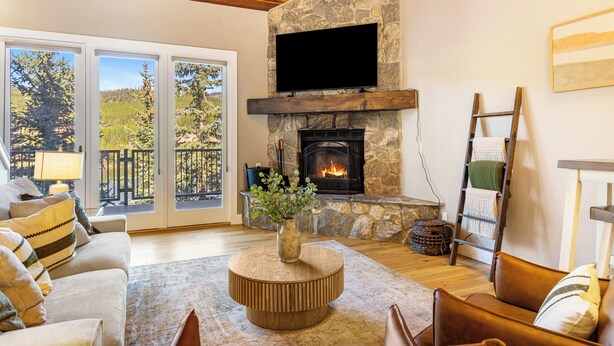 A cozy living room with a stone fireplace, a television mounted above it, and a large window overlooking a deck. - NEW! Ski-In/Out, Lone Peak & Lake Views, 3B/3B (Big Sky)