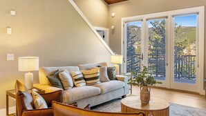 A living room with a sofa, two lamps, a coffee table, and a view of a balcony with trees. - NEW! Ski-In/Out, Lone Peak & Lake Views, 3B/3B (Big Sky)