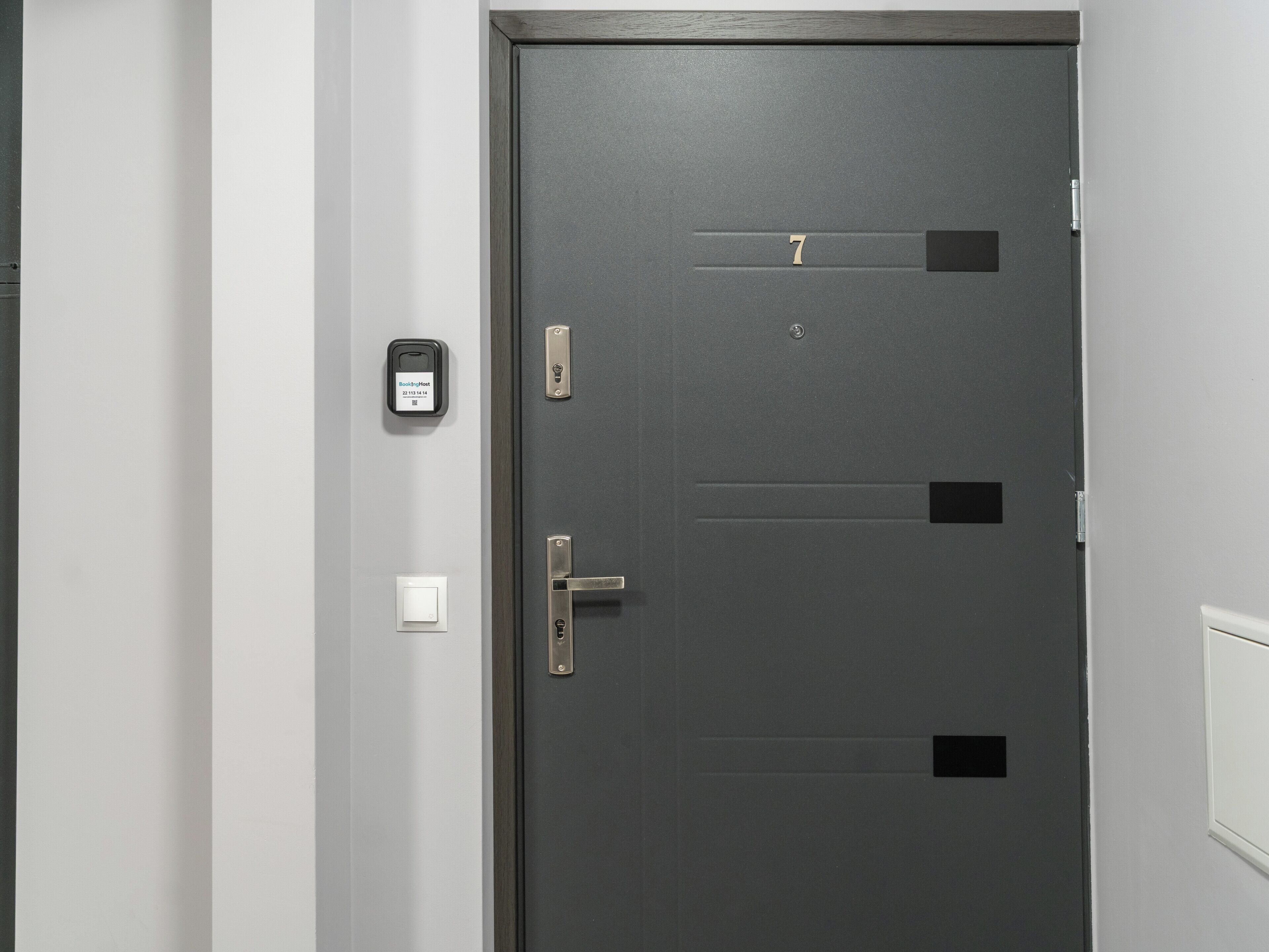 A dark grey door with a keypad lock, a doorbell, and a light switch on the wall beside it.