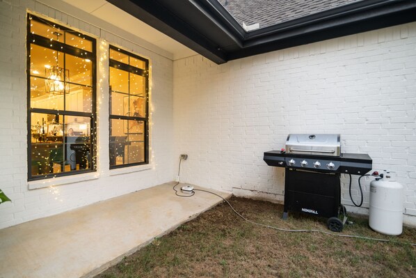 A backyard with a grill, a white brick wall, and a window with string lights.