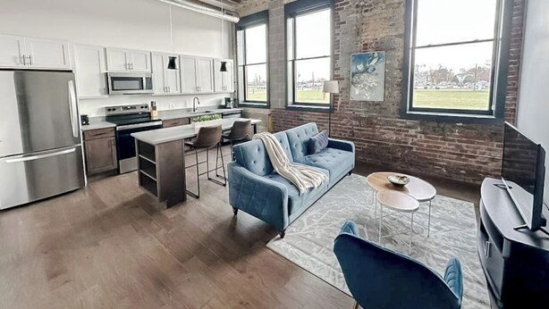 A modern kitchen with stainless steel appliances, a dining area with a blue sofa, and large windows offering a view. - Sleepover | Stylish 1BD/1BA Loft - Newton (Newton)