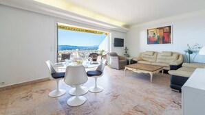A modern living room with a dining area, a sofa, a TV, and a large window with a view of the city. - 2-Br Panoramic Views – Top Floor, Cannes (Cannes)