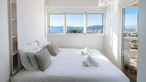 A bedroom with a large bed, white bedding, and a view of a city and mountains through a window. - 2-Br Panoramic Views – Top Floor, Cannes (Cannes)