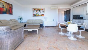 A modern living room with a beige sofa, a white side table, and a dining area with white chairs and a round table. - 2-Br Panoramic Views – Top Floor, Cannes (Cannes)