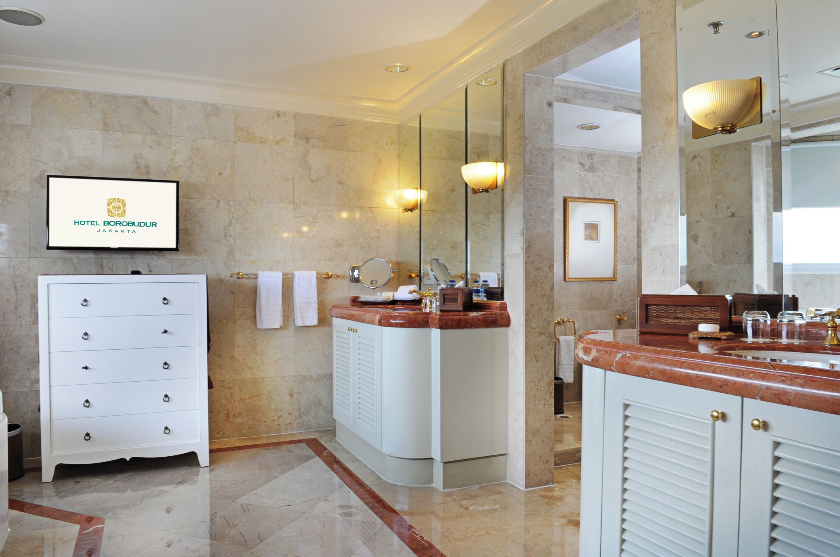 A modern bathroom with a marble floor, a white vanity with drawers, a large mirror, and a television displaying the hotel name.
