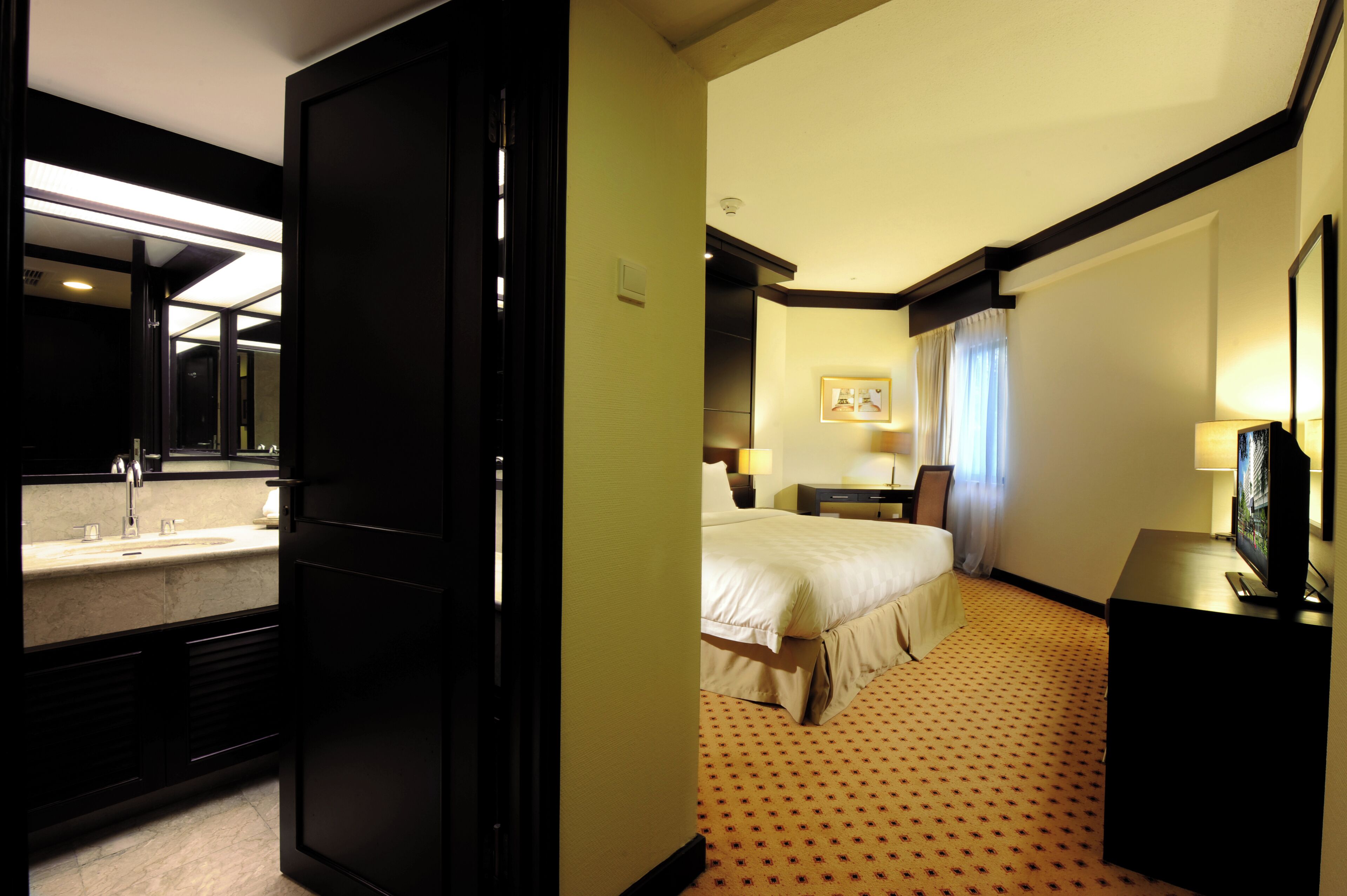 Een hotelkamer met een bed, bureau, televisie en een badkamer met wastafel en spiegel.
