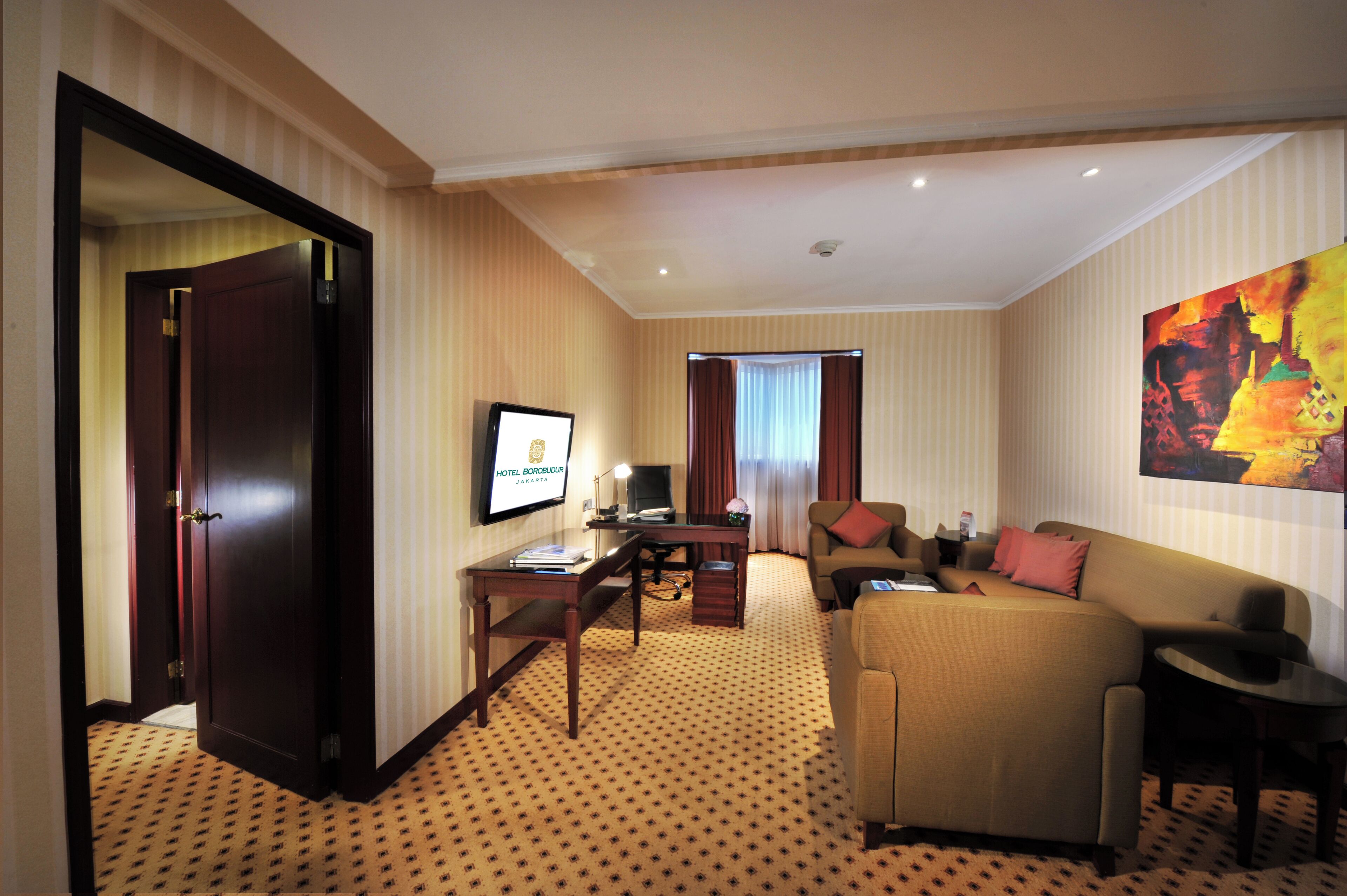 A hotel room with a living area featuring a sofa, a TV, a desk, and a carpeted floor.