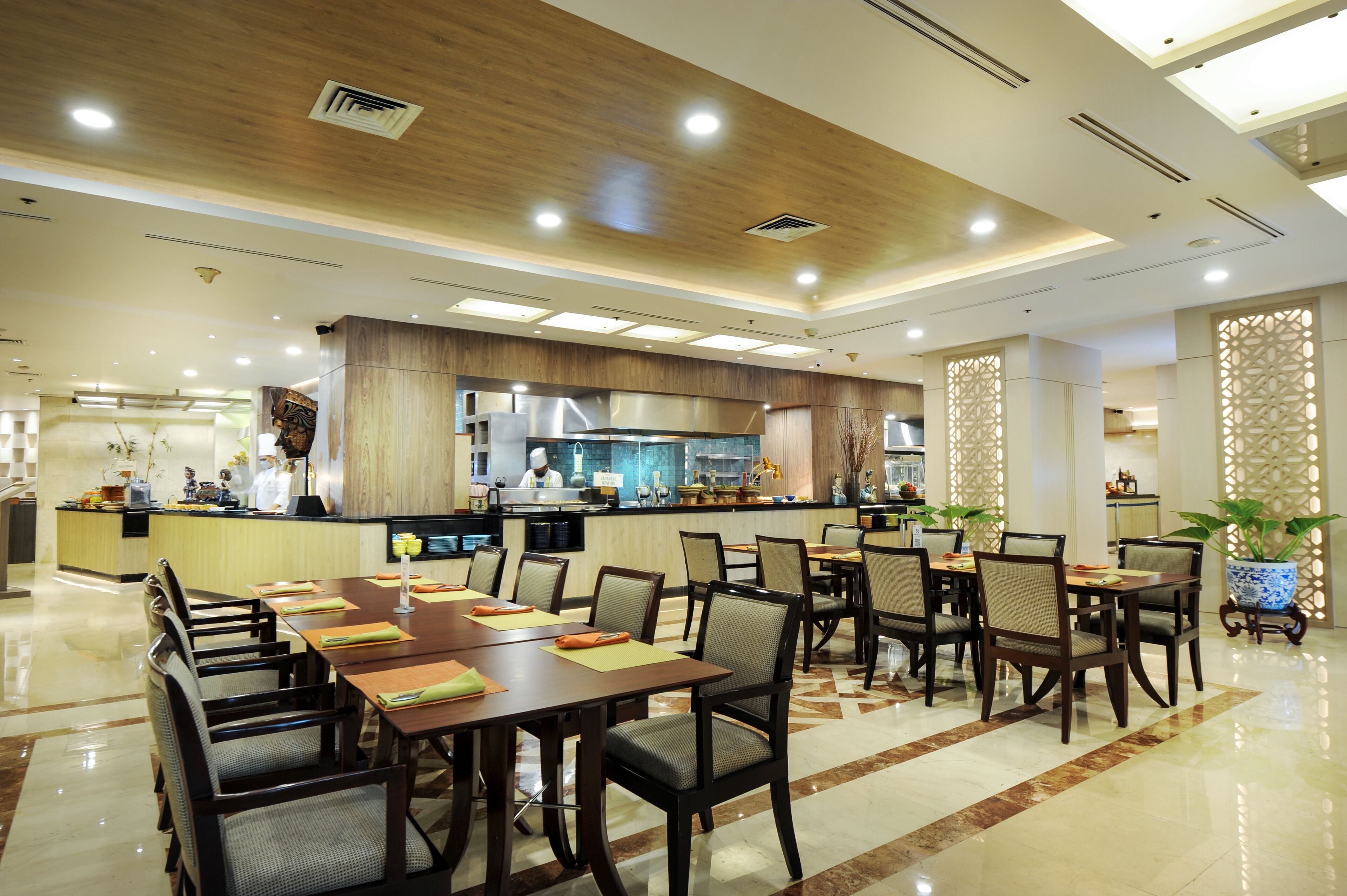 A spacious dining area with multiple tables and chairs, a central kitchen area, and decorative plants.