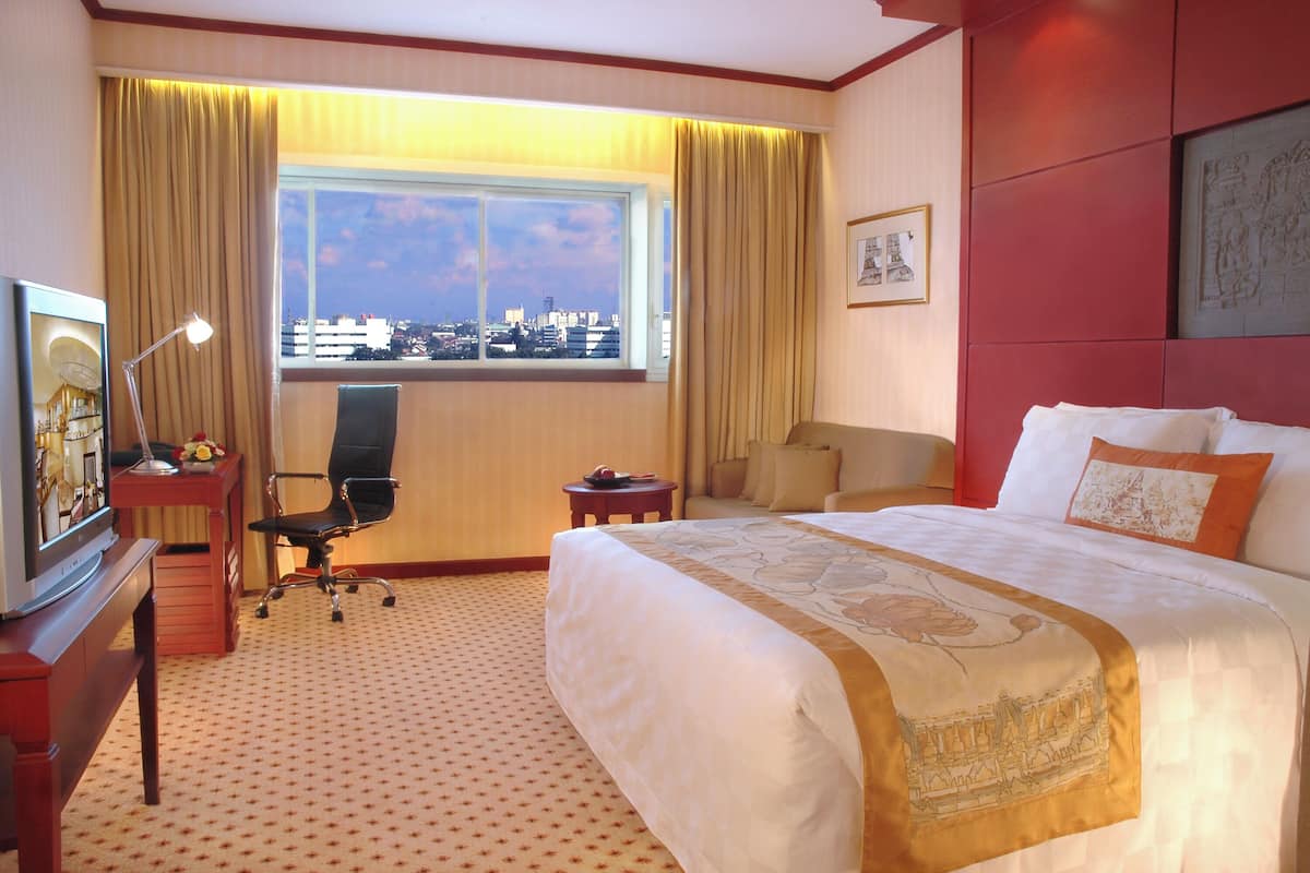 Habitación de hotel con una cama grande, un escritorio con lámpara, una silla, una mesita y una ventana con vistas a la ciudad.