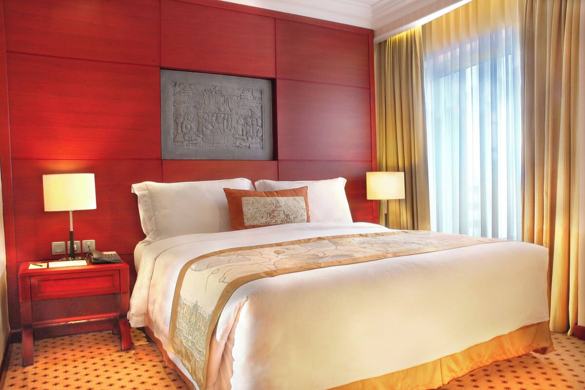 Habitación de hotel con una cama grande, dos mesitas de noche, una lámpara de noche, una obra de arte en la pared y una ventana con cortinas.
