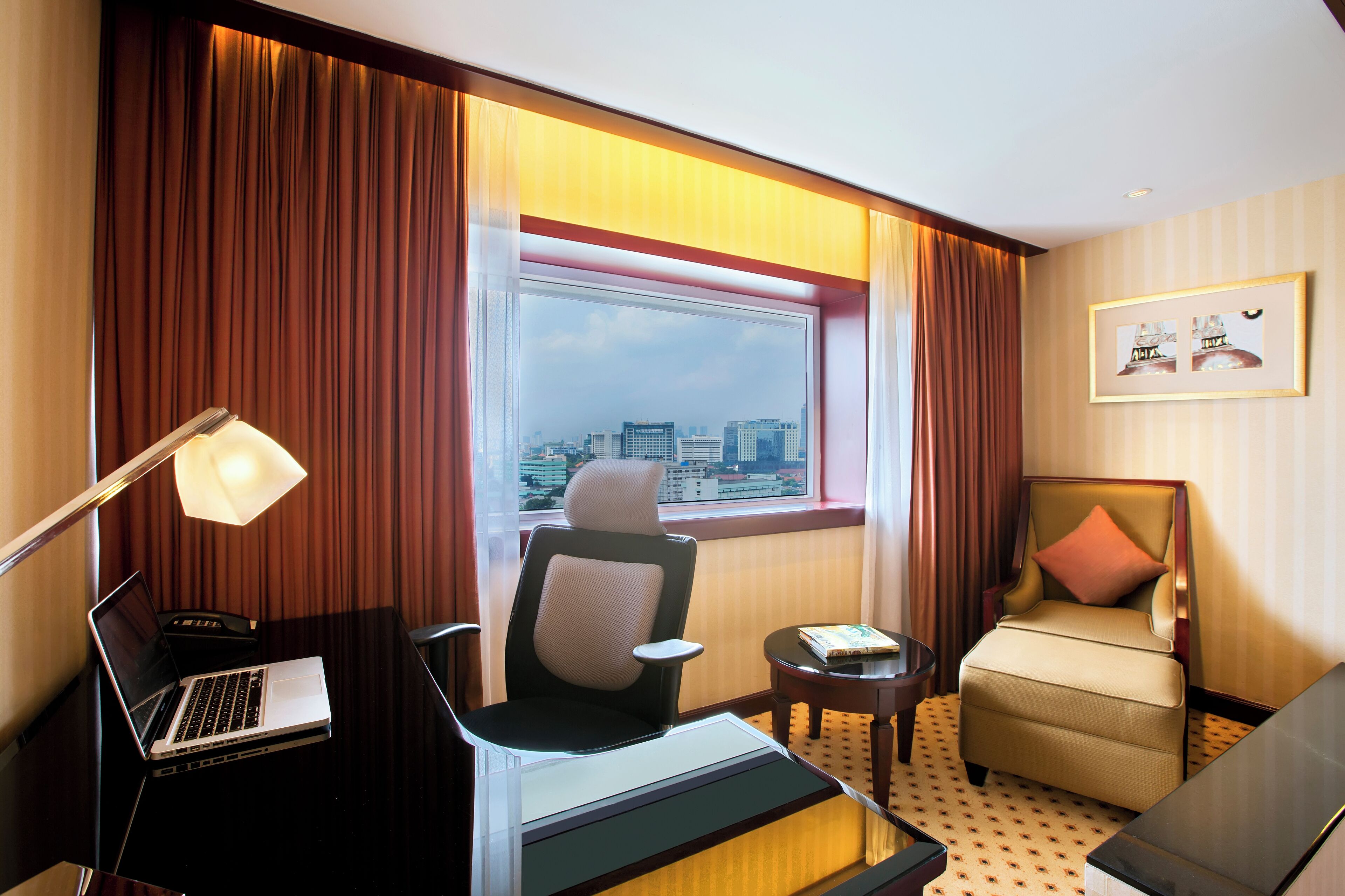 A hotel room with a city view, a desk with a laptop, a comfortable armchair, and a small side table.