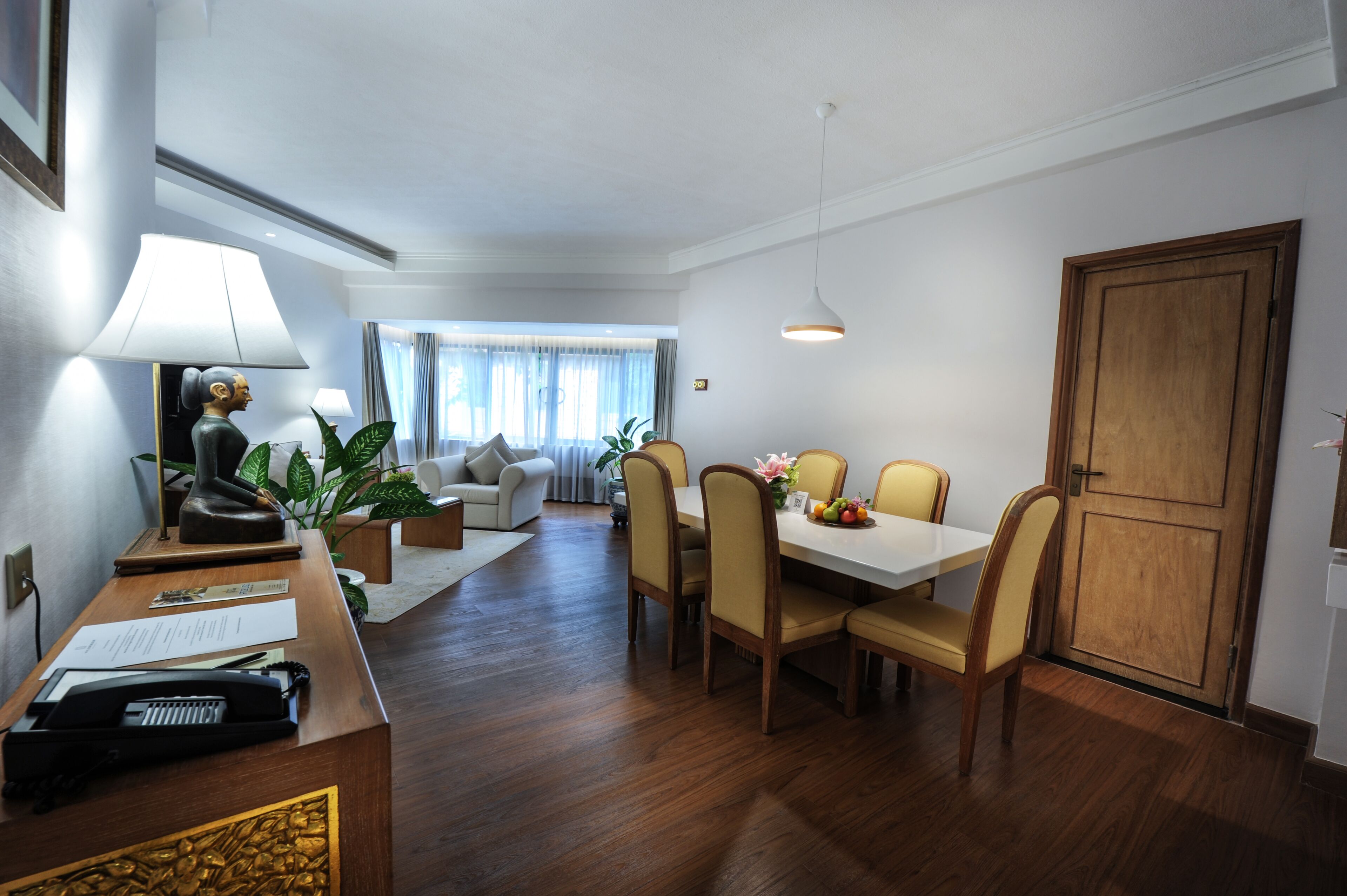 A hotel room with a dining area, a sofa, a desk with a lamp, and a wooden door.