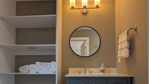 A bathroom with a dark blue vanity, a round mirror, and shelves with folded towels. - Modern Raleigh Downtown Rooftop Skyline View SFH-R (Raleigh)