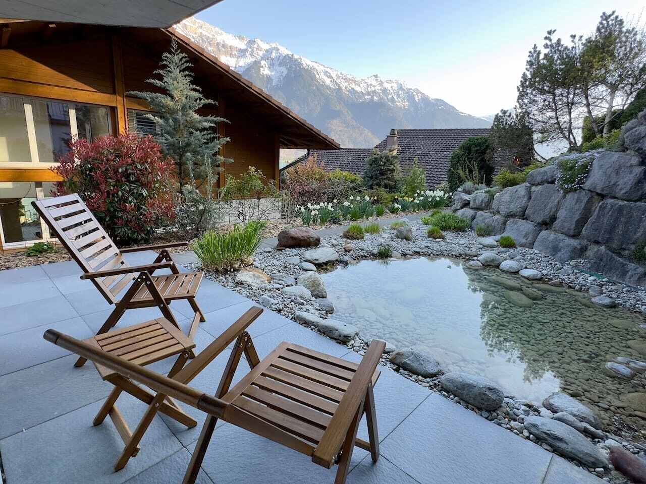 Terraza de madera con sillones, un estanque pequeño y montañas al fondo.