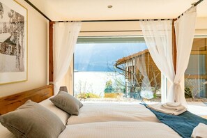 A bedroom with a large bed, white curtains, and a view of a wooden structure and mountains.
