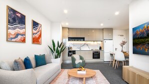 A modern living room with a sofa, coffee table, and artwork. A kitchen is visible in the background with cabinets and appliances. - Modern City Views Escape With Gym Access (Melbourne)