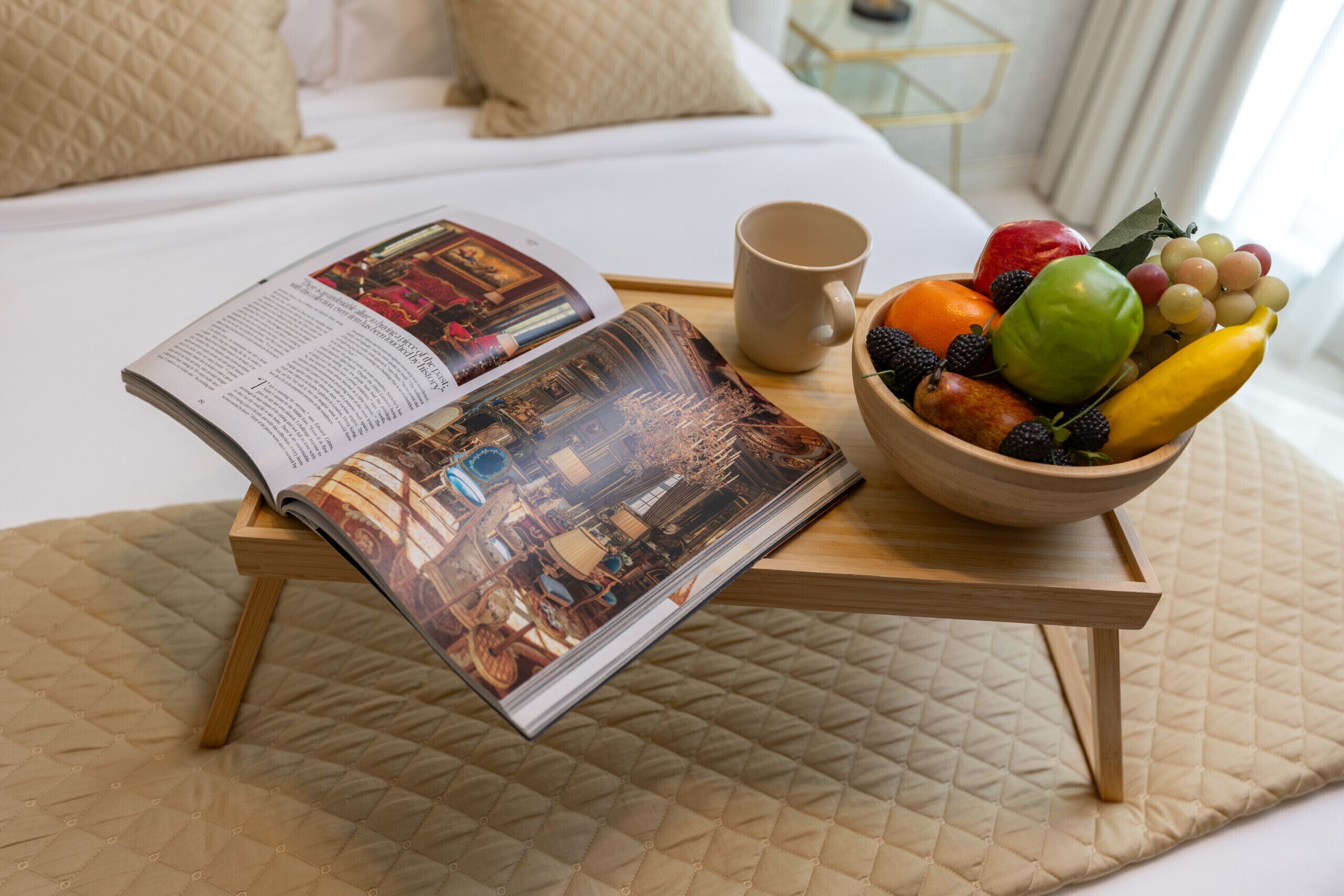 Ein Bett mit gesteppter Bettdecke, eine Zeitschrift auf einem Holztisch, eine Obstschale und eine Tasse auf einem Tablett.